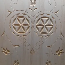 Jasne, surowe drewno z wyżłobionym wzorem w kształcie serca, zawierającym dwa okręgi z geometrycznym motywem kwiatowym oraz stylizowane motywy roślinne.