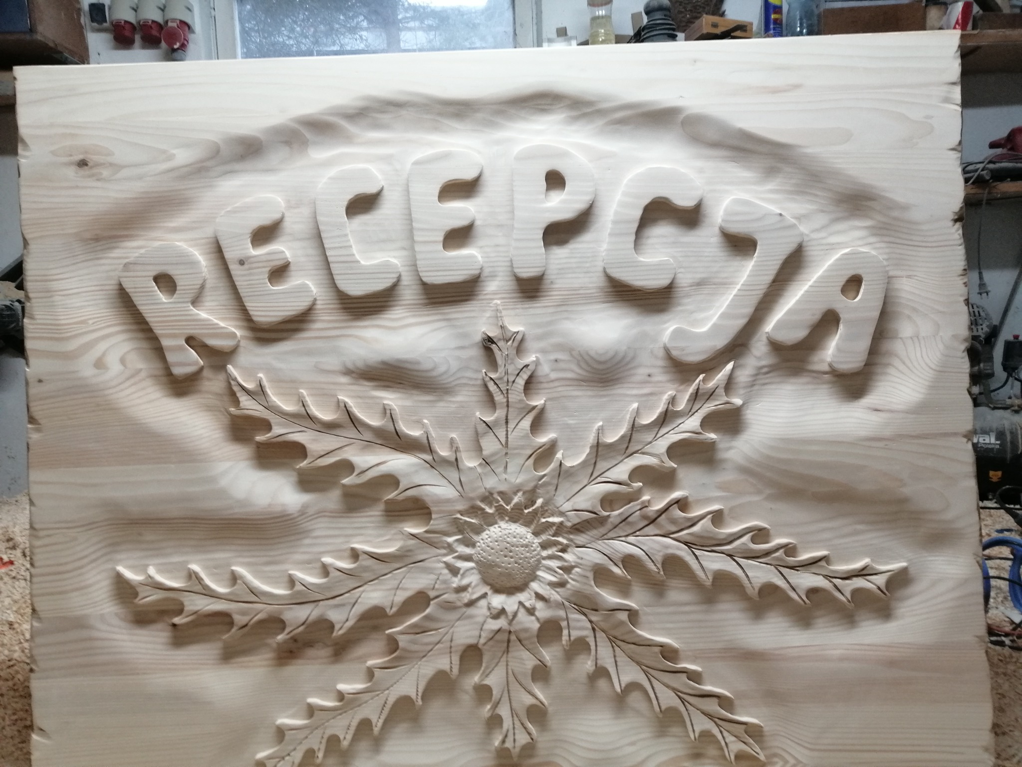 Drewniana tablica z wyżłobionym napisem 'RECEPCJA' i motywem roślinnym z liśćmi dębu, surowe drewno w warsztacie stolarskim.