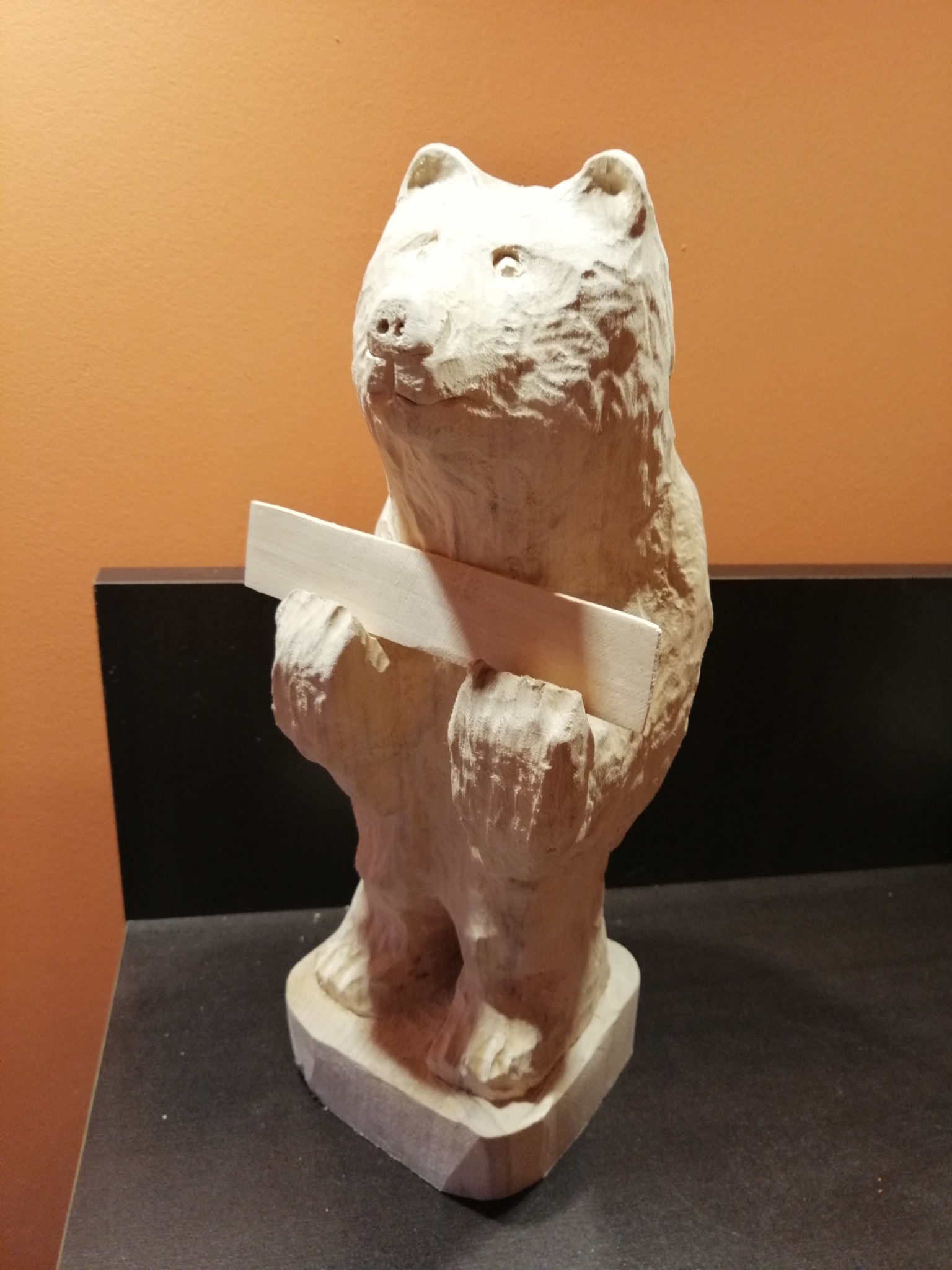 Niedokończona, surowa rzeźba w drewnie przedstawiająca stojącego misia trzymającego prostokątny kawałek drewna, widoczne ślady dłuta i obróbki