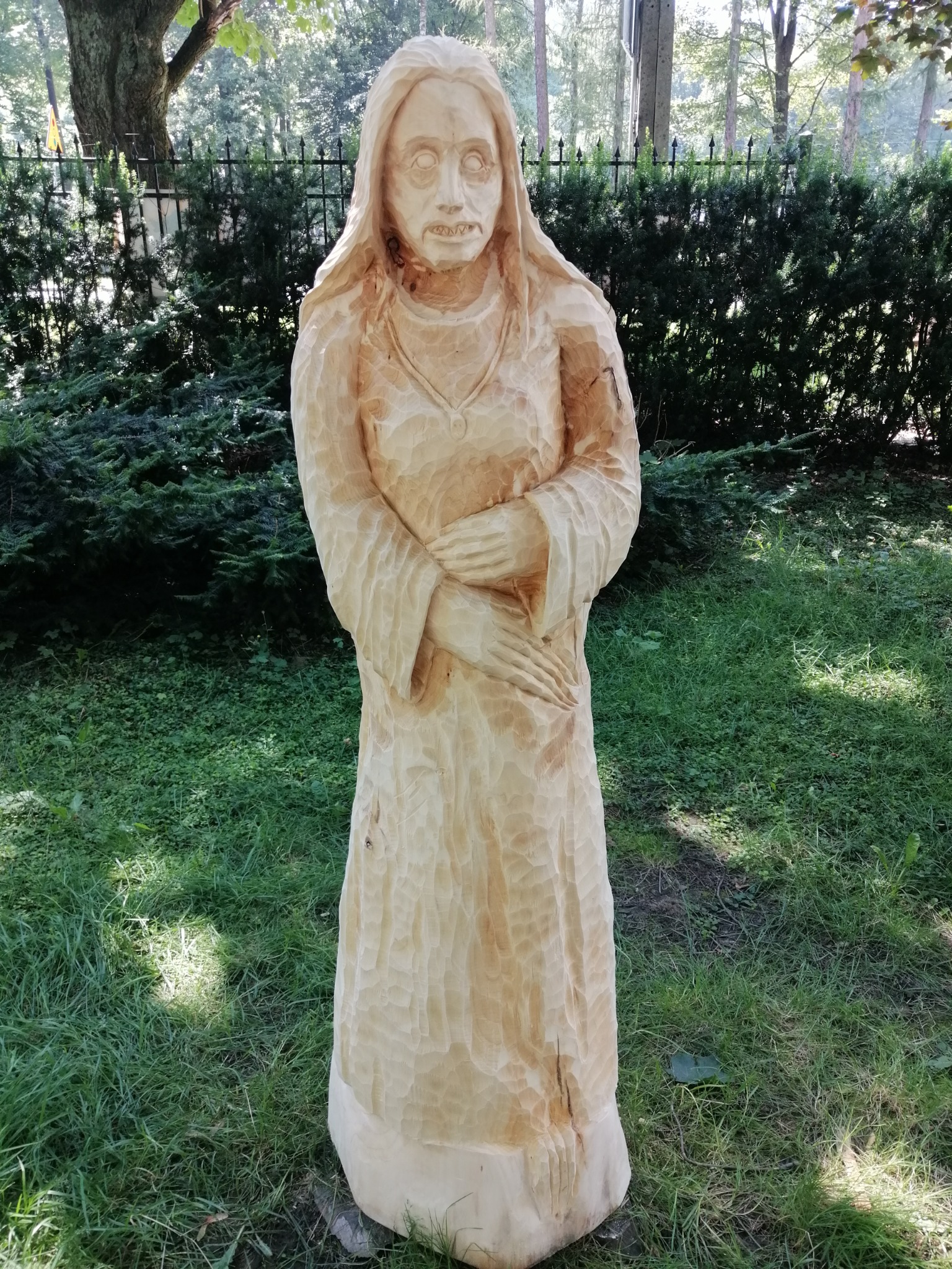 Drewniana rzeźba kobiety o surowym wykończeniu, ustawiona na trawniku w parku, widoczne ślady dłuta.
