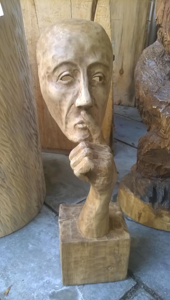 Drewniana rzeźba przedstawiająca popiersie z palcem wskazującym na ustach, umieszczona na kwadratowej podstawie, obok innych drewnianych rzeźb o nieokreślonych kształtach.