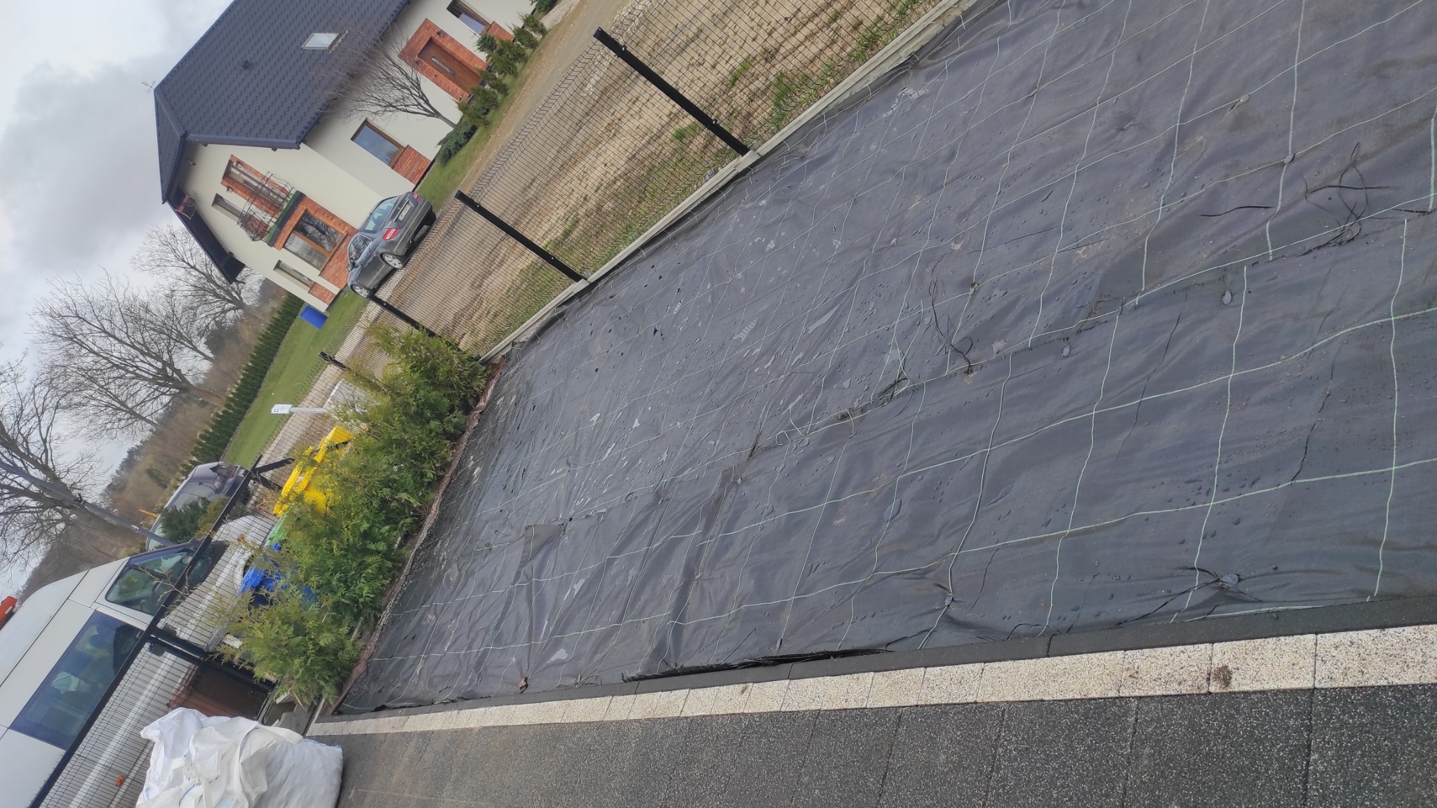 Rozłożona czarna agrowłóknina z zieloną siatką na przygotowanym podłożu, widoczne kołki mocujące, w tle rząd tuj i dom jednorodzinny.