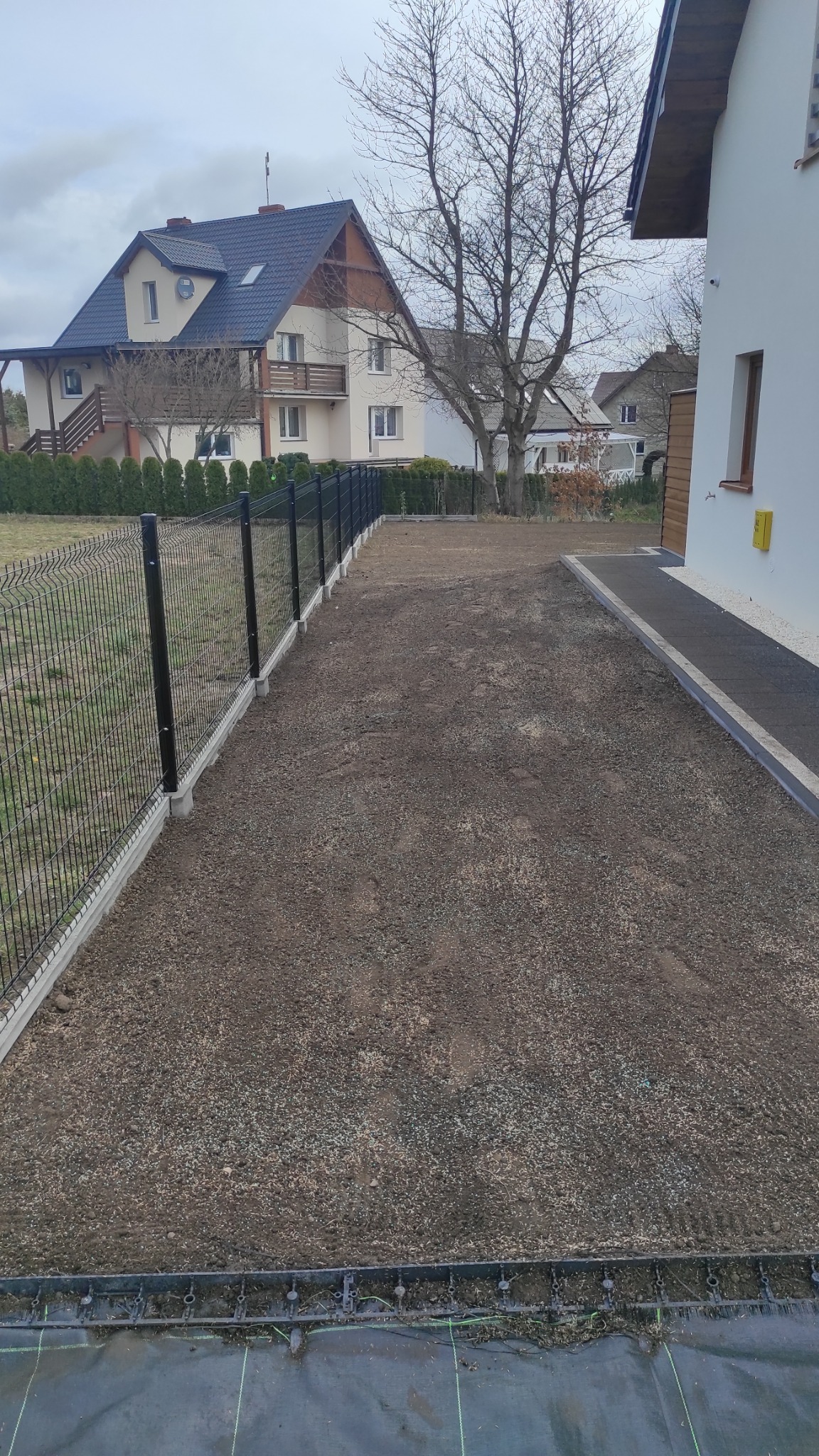 Przygotowany teren pod trawnik z widocznymi nasionami i siatką zabezpieczającą, obok nowoczesny dom z białymi ścianami i czarnym dachem, w tle dom jednorodzinny.