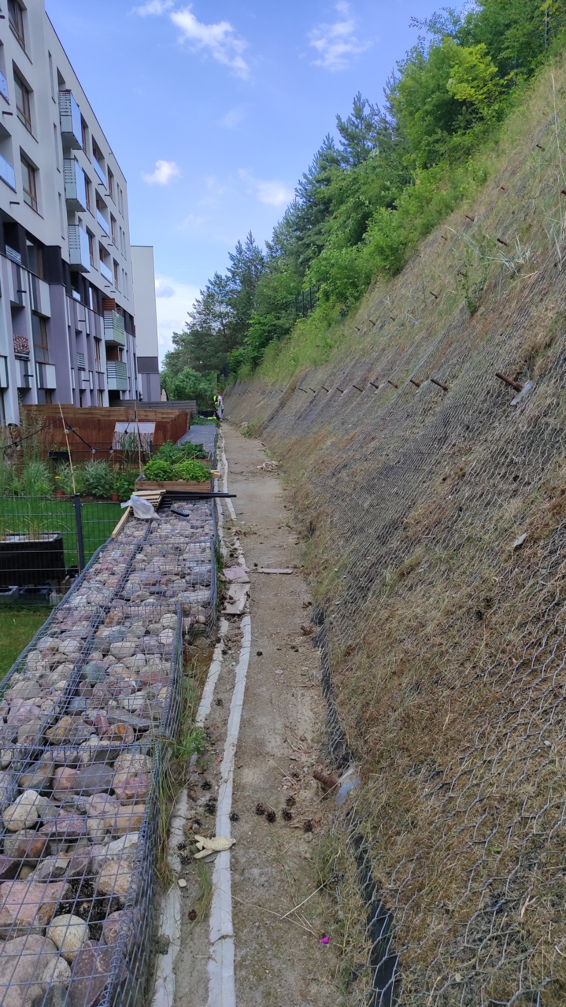 Skarpa zabezpieczona siatką geotechniczną obok budynku mieszkalnego, wzdłuż chodnika z gabionami wypełnionymi kamieniami. W tle widoczna osoba w kamizelce odblaskowej.