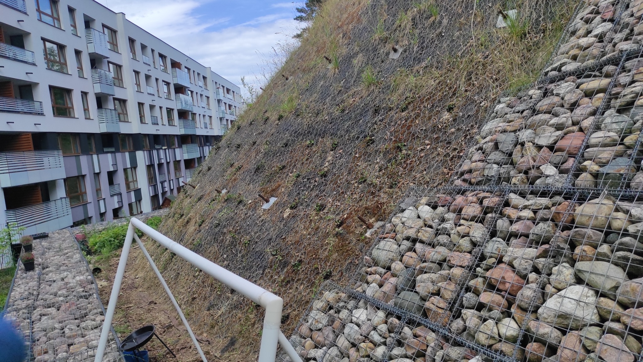 Skarpa zabezpieczona siatką i kamieniami gabionowymi, widok z dołu na tle nowoczesnego budynku wielorodzinnego.
