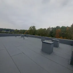 Szary, płaski dach pokryty papą termozgrzewalną z trzema kominami wentylacyjnymi i przewodami instalacji na tle lasu i pochmurnego nieba.