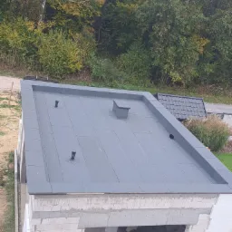 Płaski dach pokryty szarą papą termozgrzewalną, widoczne kominki wentylacyjne i obróbki blacharskie, budynek w trakcie budowy, w tle zieleń drzew i dach sąsiedniego budynku.