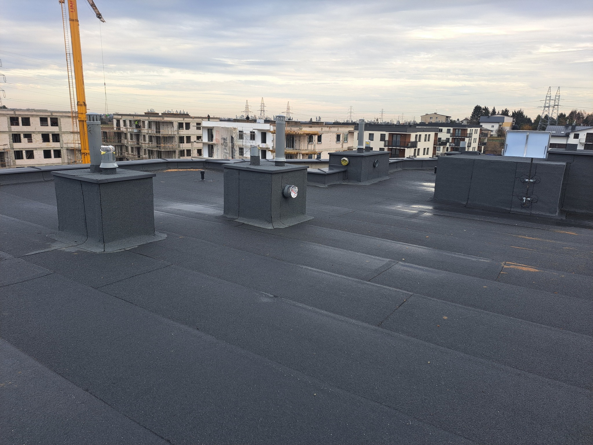 Czarny, płaski dach pokryty papą termozgrzewalną z kominami wentylacyjnymi na tle budynków w trakcie budowy i dźwigu.