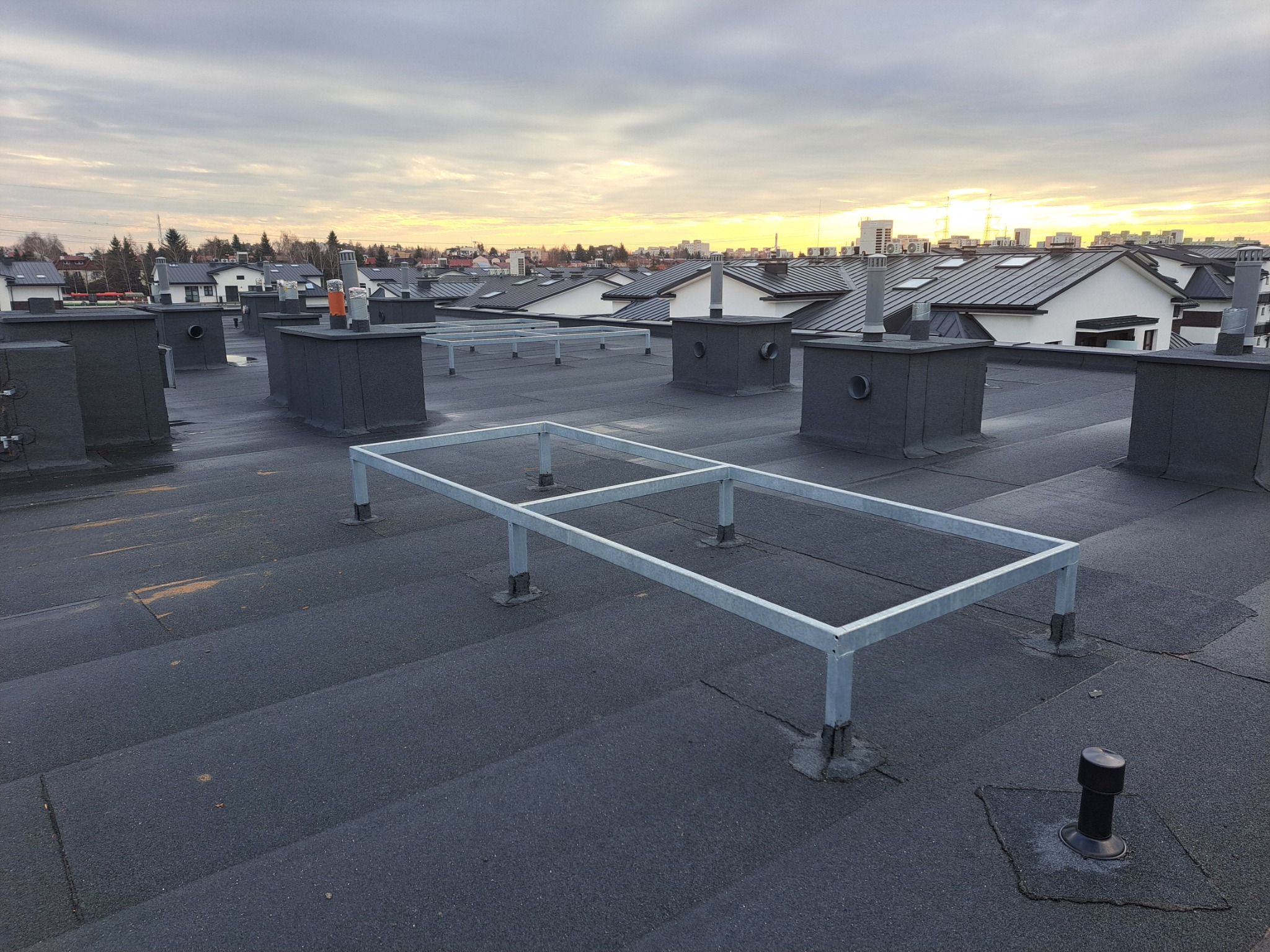 Widok na płaski dach pokryty papą z wentylacją i metalową konstrukcją wsporczą, w tle zabudowa jednorodzinna i zachodzące słońce.