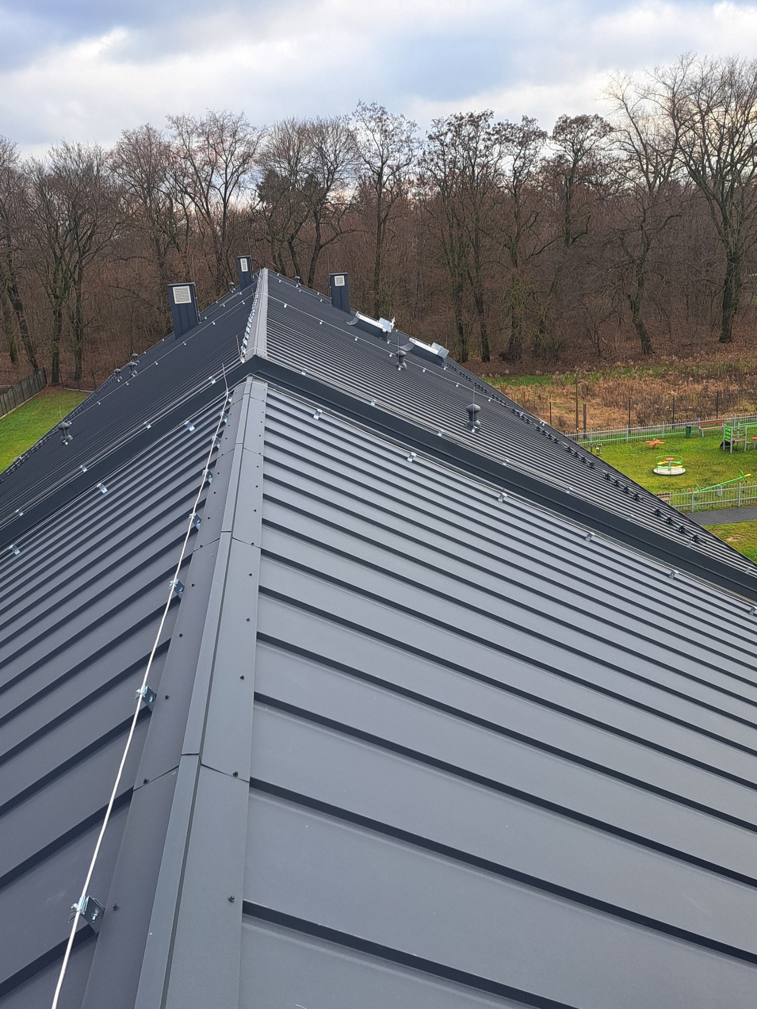 Nowoczesny, grafitowy dach panelowy z systemem odgromowym, widok z góry na tle drzew i placu zabaw.