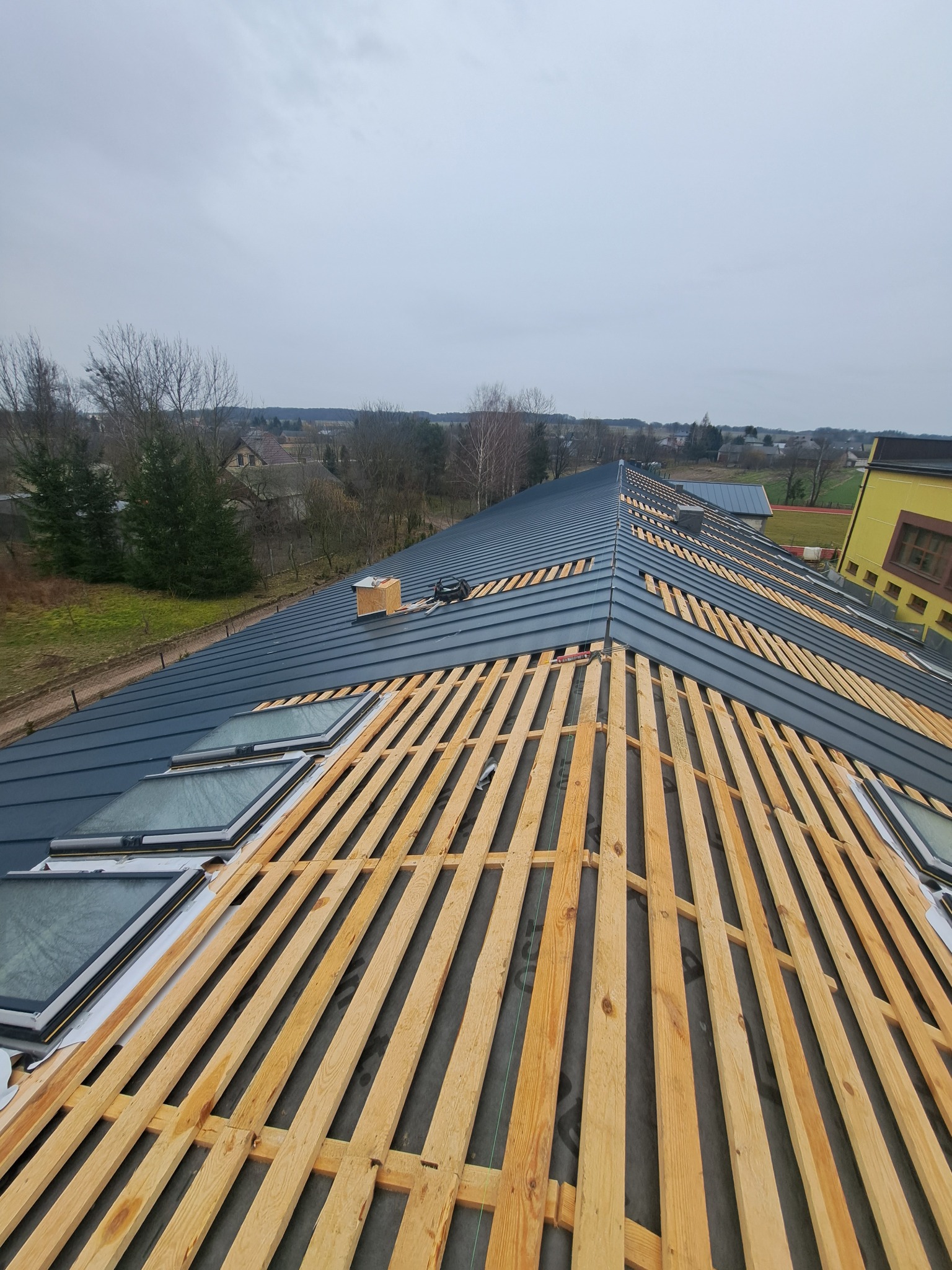 Widok z góry na konstrukcję dachu z zamontowanymi oknami dachowymi oraz drewnianą łatą, przygotowaną pod pokrycie blachodachówką w kolorze grafitowym. W tle widoczny krajobraz wiejski i pochmurne...