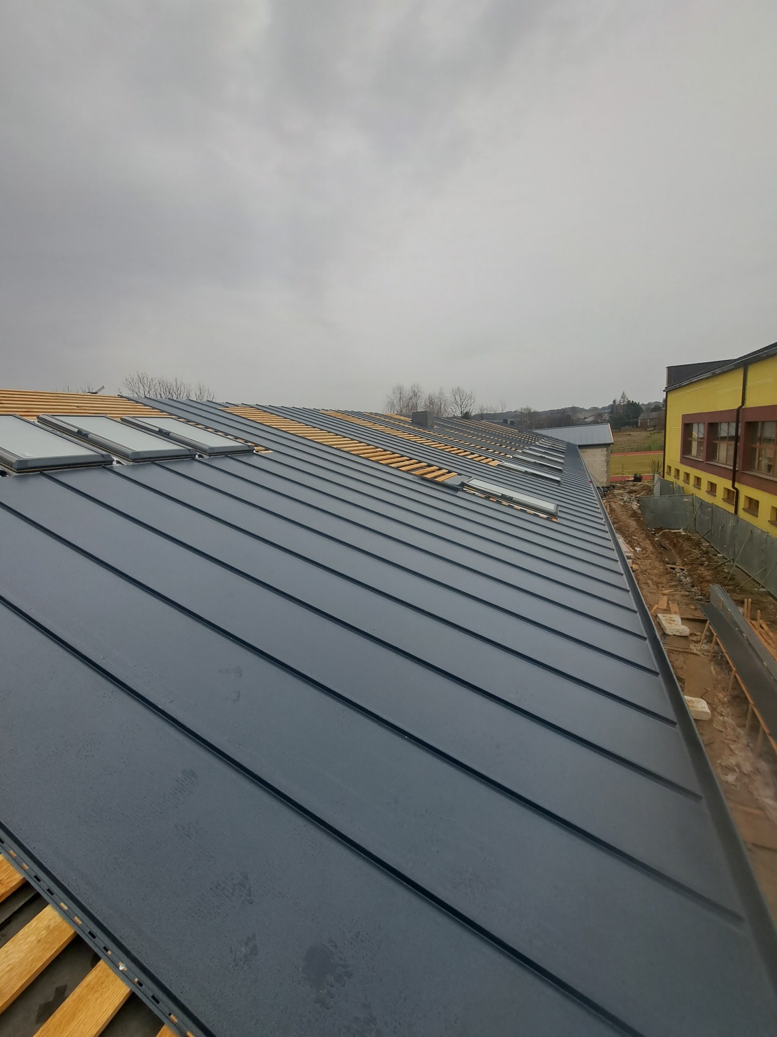 Nowy, grafitowy dach z blachy na budynku, z zamontowanymi oknami dachowymi i widoczną częścią drewnianej konstrukcji pod spodem, na tle pochmurnego nieba i części budynku w kolorze żółtym.