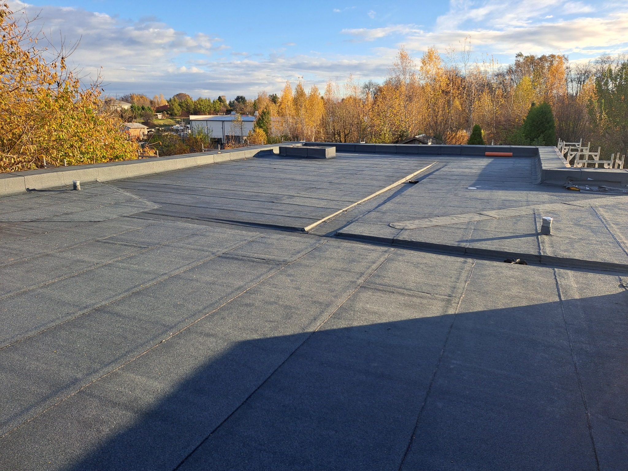 Widok z góry na płaski dach pokryty papą termozgrzewalną w jesiennym krajobrazie, widoczne kominki wentylacyjne i elementy wykończeniowe.