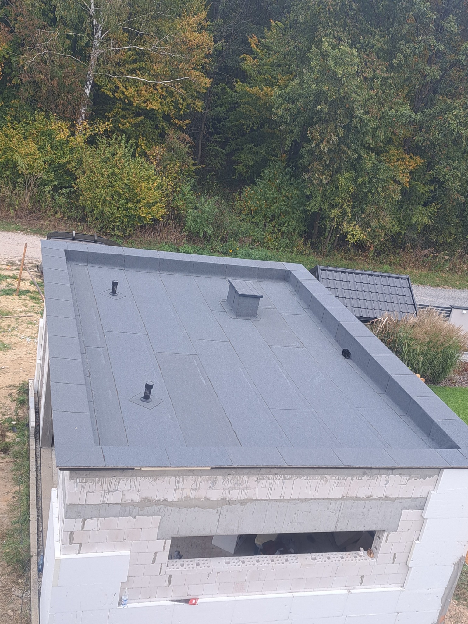Płaski dach pokryty szarą papą termozgrzewalną, widoczne kominki wentylacyjne i obróbki blacharskie, budynek w trakcie budowy, w tle zieleń drzew i dach sąsiedniego budynku.