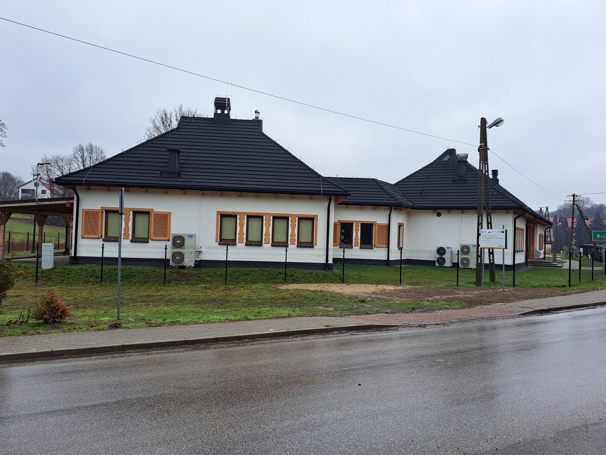 Budynek z nowym, ciemnym dachem z dachówki ceramicznej, widoczne kominy i obróbki blacharskie, biała elewacja z drewnianymi okiennicami, pochmurny dzień.