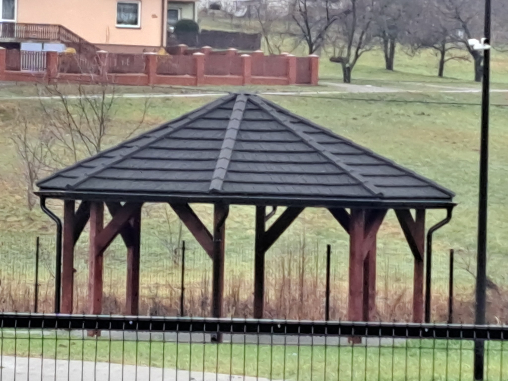 Zadaszona altana z ciemną dachówką ceramiczną, drewnianą konstrukcją i rynnami, widoczna zza metalowego ogrodzenia na tle zielonego wzgórza i zabudowań.