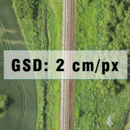 Zdjęcie z drona przedstawia linię kolejową przecinającą zielony krajobraz z polami i lasami, widok z góry.