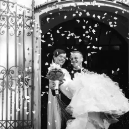 Czarno-białe zdjęcie uśmiechniętej pary młodej wychodzącej z kościoła, obsypywanej konfetti w kształcie płatków, widoczne ozdobne, metalowe drzwi.