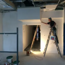 Mężczyzna na drabinie maluje pędzlem sufit w pomieszczeniu w trakcie prac wykończeniowych, widoczny ruszt budowlany, puszki z farbą i przewody elektryczne.