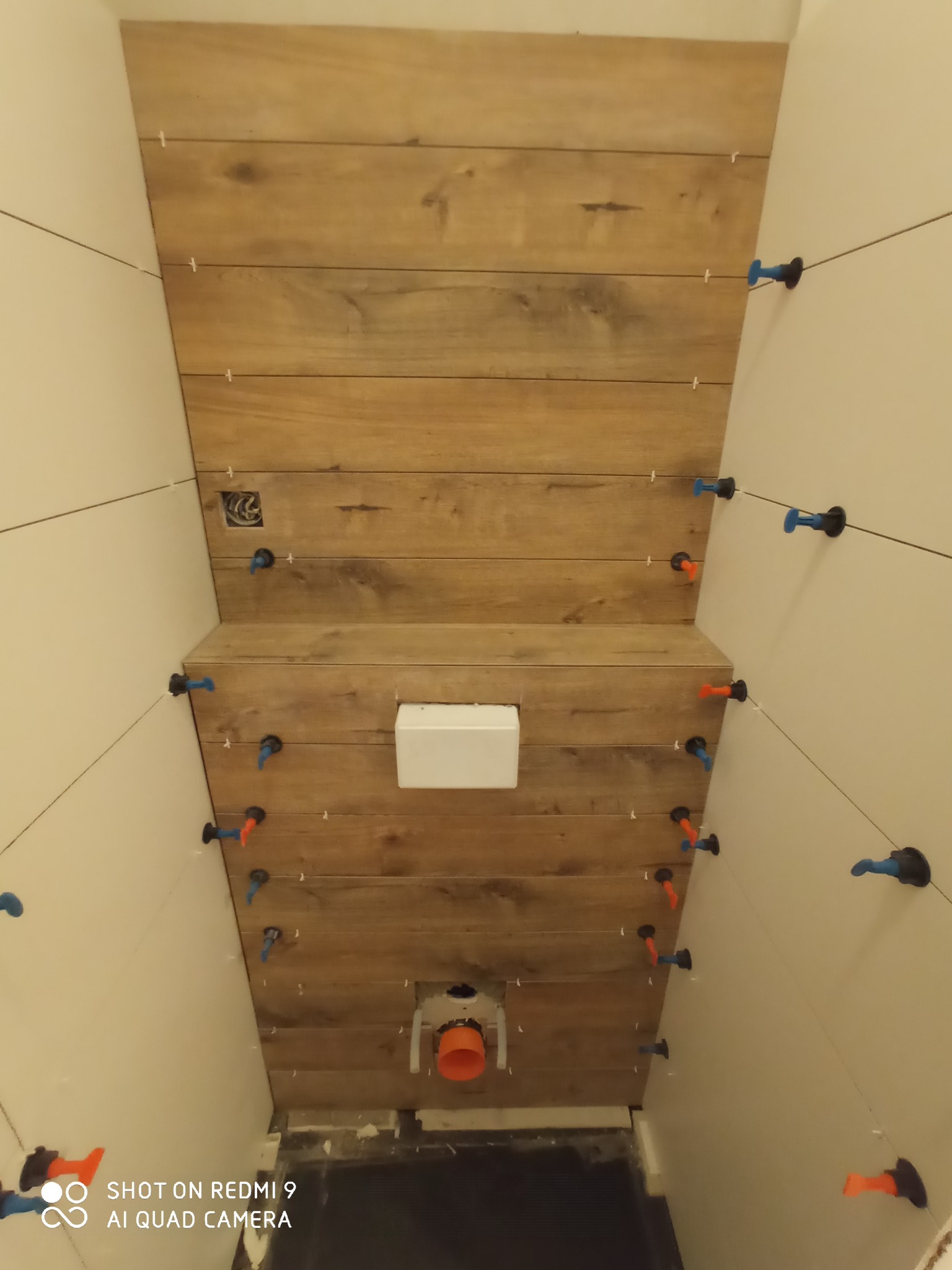 Ściana łazienki w trakcie układania płytek imitujących drewno, widoczne systemy poziomowania płytek i instalacja podtynkowego WC.