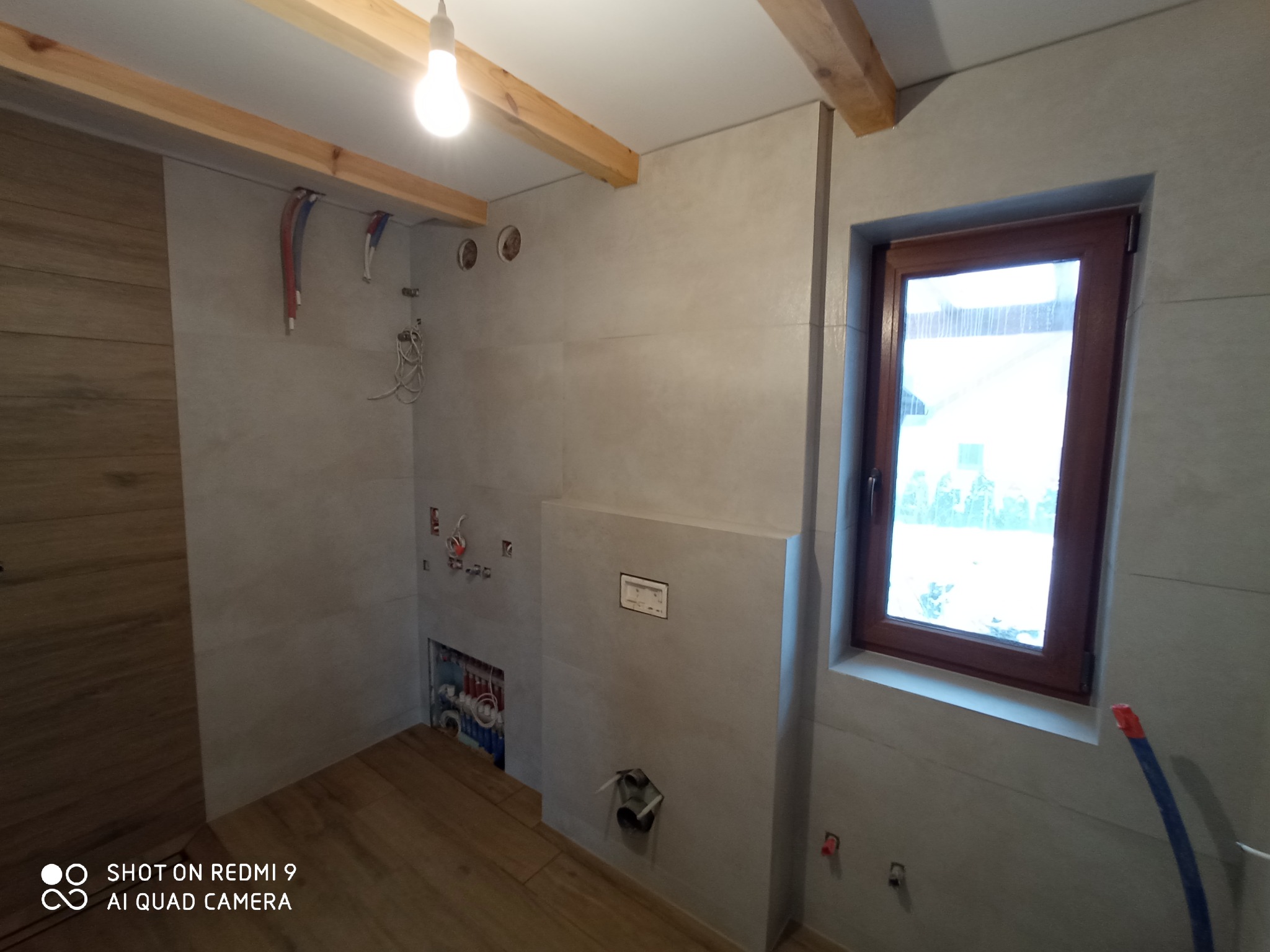 Niedokończona łazienka z szarymi płytkami, drewnianymi belkami stropowymi, oknem z brązową ramą i widoczną instalacją hydrauliczną w ścianie.
