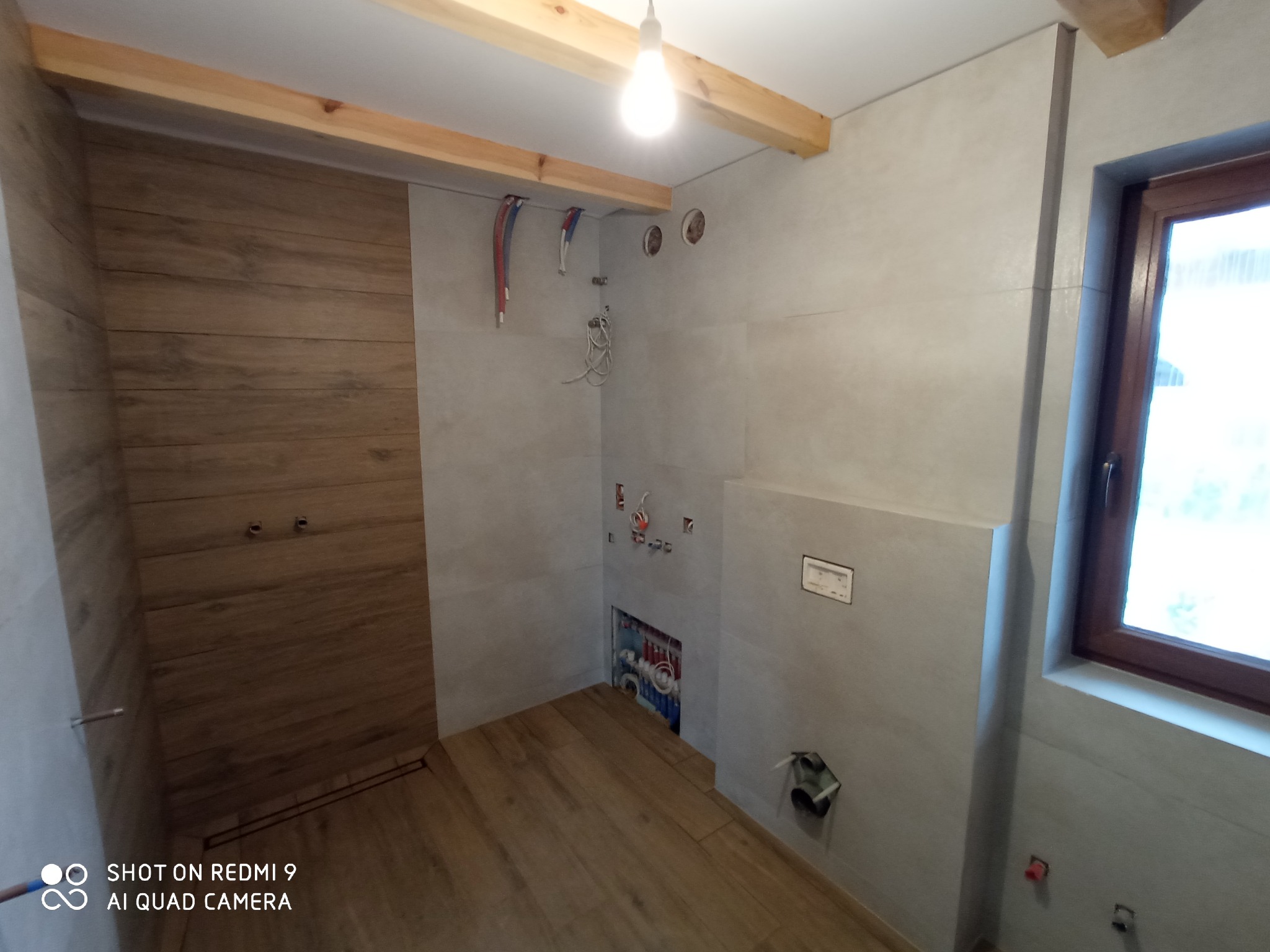 Wnętrze łazienki w trakcie prac wykończeniowych: położone płytki imitujące drewno na podłodze i ścianie w strefie prysznica, szare płytki na pozostałych ścianach, widoczne instalacje...