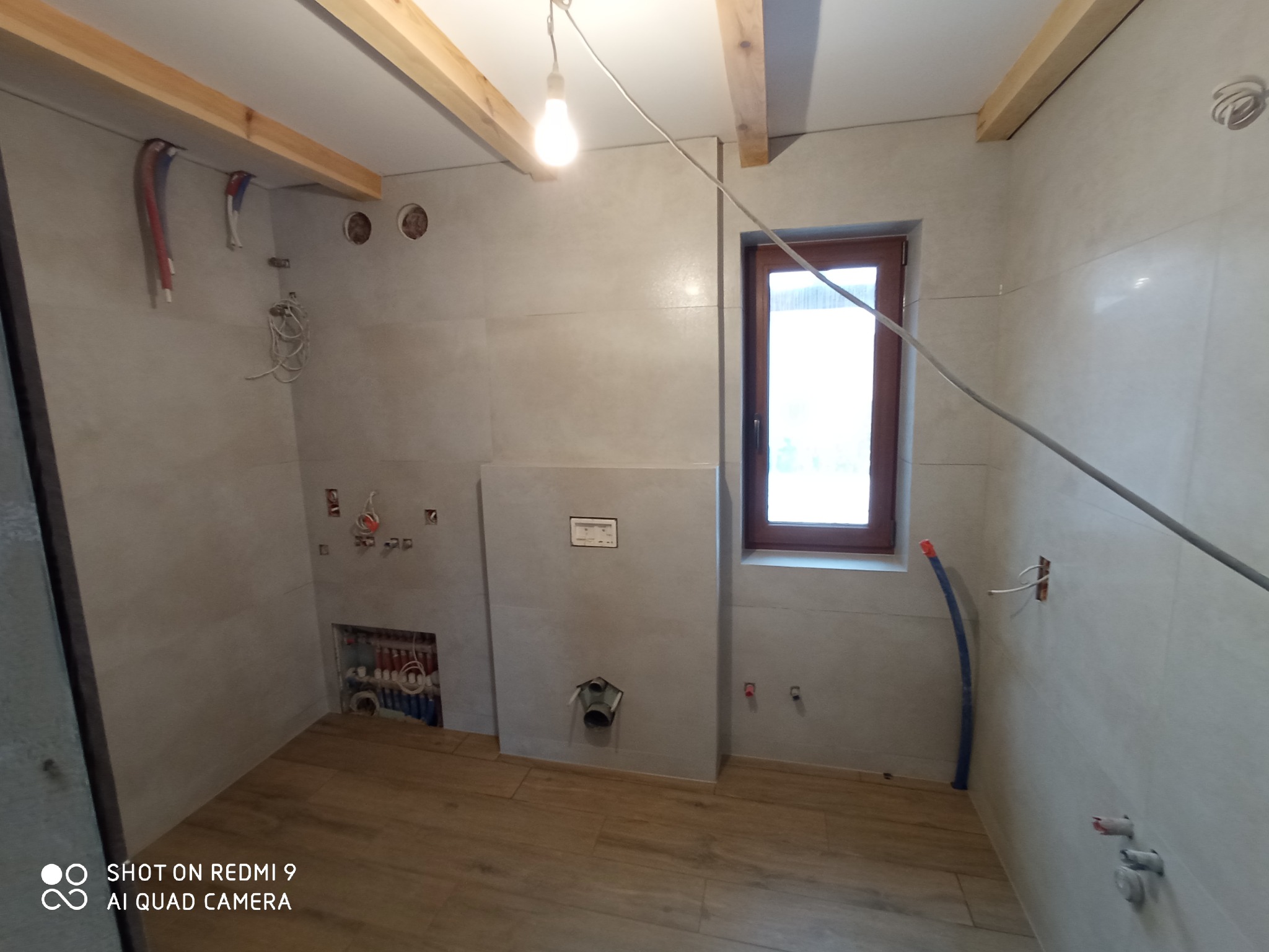 Wnętrze łazienki w trakcie prac wykończeniowych: ułożone płytki ceramiczne na ścianach i podłodze imitujące drewno, widoczne instalacje hydrauliczne i elektryczne, surowe drewniane belki stropowe...