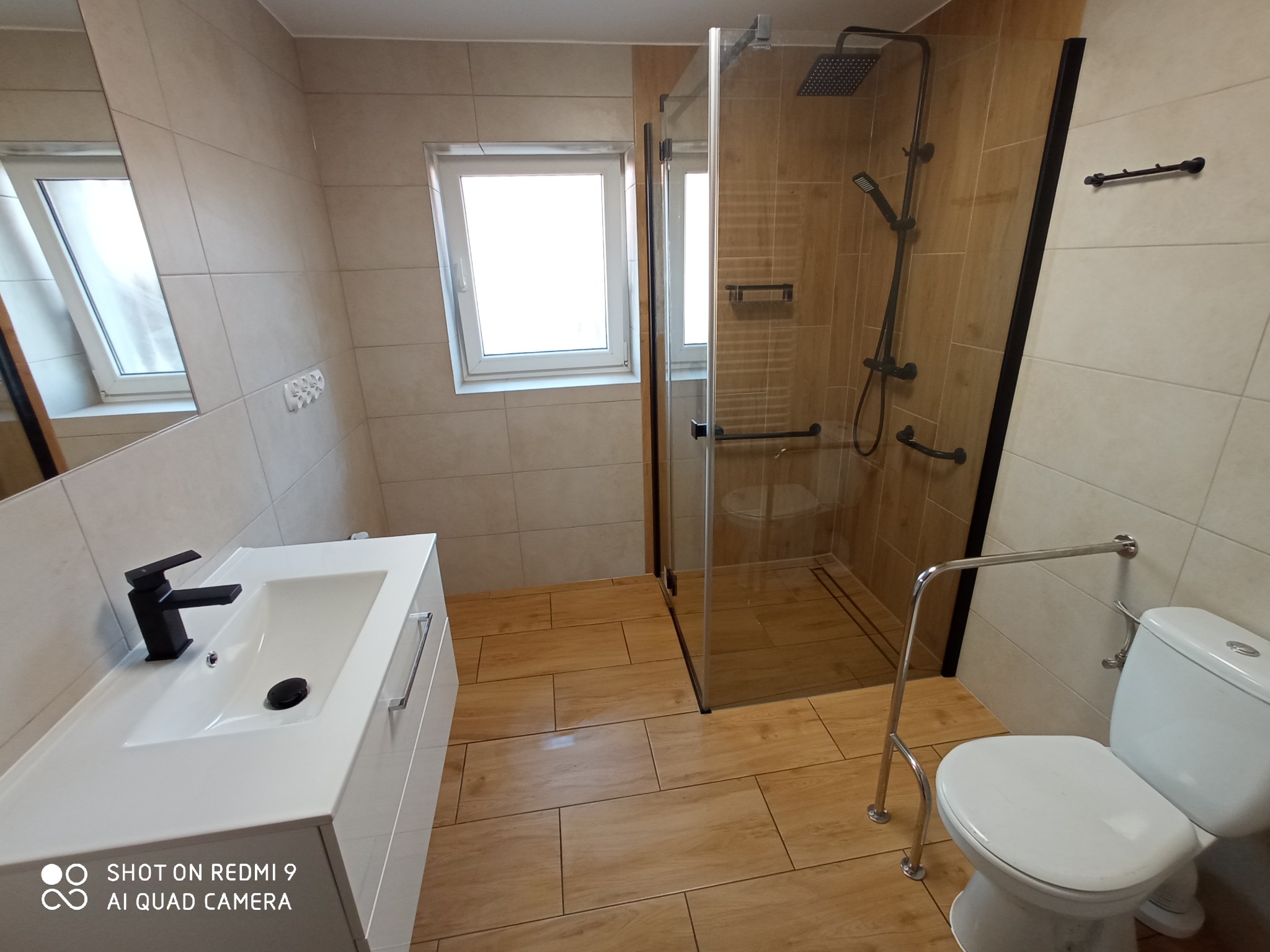 Jasna łazienka z kabiną prysznicową typu walk-in z czarnymi elementami, białą umywalką z czarną baterią i toaletą z uchwytem dla osoby niepełnosprawnej.