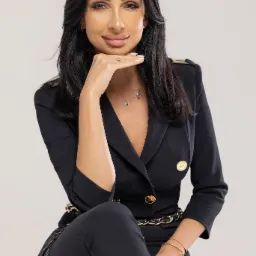 Portret kobiety w czarnym, eleganckim garniturze z ozdobnymi guzikami i złotym paskiem, opierającej brodę na dłoni z pierścionkiem, na jasnym tle.