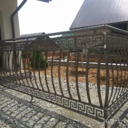 Metalowa konstrukcja balustrady z motywem greckim, surowy stan, widoczna na żwirowym podłożu w trakcie montażu, w tle budynek z brązowym dachem.