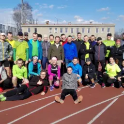 Grupa biegaczy pozuje do zdjęcia na bieżni lekkoatletycznej w słoneczny dzień, widoczny budynek w tle.