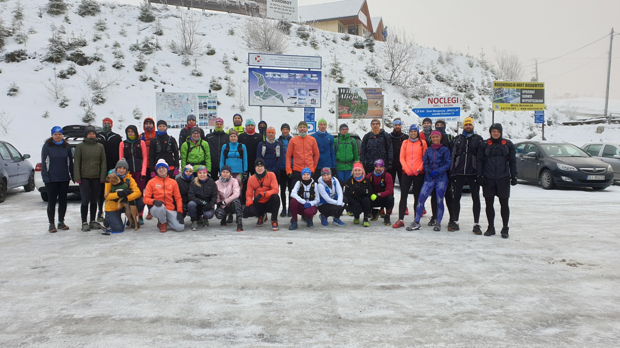Grupa biegaczy w zimowym krajobrazie, pozująca do zdjęcia po treningu, widoczny śnieg i tablice informacyjne w tle, jedna osoba trzyma psa.