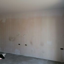 KPT Piotr - Ściana po szpachlowaniu, widoczne gniazdka elektryczne i przewody, przygotowana do dalszych prac wykończeniowych, z materiałami budowlanymi na podłodze.