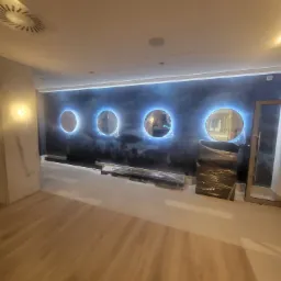 Ściana z efektem ombre w odcieniach granatu i szarości, ozdobiona czterema podświetlanymi okrągłymi dekoracjami, częściowo zabezpieczona folią ochronną, w trakcie prac wykończeniowych.