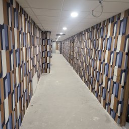 KPT Piotr - Długi korytarz wykończony tapetą w geometryczne wzory w odcieniach brązu, bieli i błękitu, widok z perspektywy posadzki w trakcie prac wykończeniowych, z widocznymi śladami po budowie.