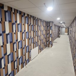 KPT Piotr - Długi korytarz biurowy z nowo położoną tapetą w geometryczne wzory w odcieniach brązu, błękitu i bieli, sufit podwieszany z widocznymi przewodami. Podłoga surowa, w trakcie wykończenia.
