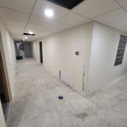 KPT Piotr - Długi korytarz w trakcie remontu z surowymi ścianami przygotowanymi pod malowanie, widoczny fragment instalacji elektrycznej, sufit podwieszany z otworami, betonowa posadzka z plamami, mała biała...