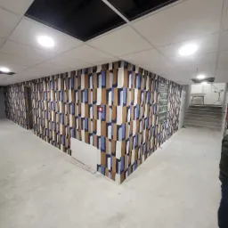 Korytarz z nową tapetą w geometryczne wzory w odcieniach brązu i błękitu, wykończony sufit z panelami i oświetleniem punktowym, w trakcie remontu.