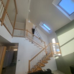 KPT Piotr - Malowanie ścian w dwupoziomowym domu ze schodami. Pracownik na drabinie maluje ścianę w pobliżu drewnianej balustrady. Widoczne okna dachowe i lampy wiszące.