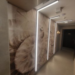 KPT Piotr - Korytarz z tapetą w beżowe wzory, oświetlony liniowymi lampami LED. Na ścianie widoczne okrągłe włączniki. W tle drzwi i dekoracyjna lampa ścienna.