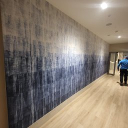 KPT Piotr - Długa ściana pokryta tapetą z abstrakcyjnym wzorem w odcieniach bieli i granatu, imitującym pionowe smugi. W tle widoczne drzwi i fragment osoby.