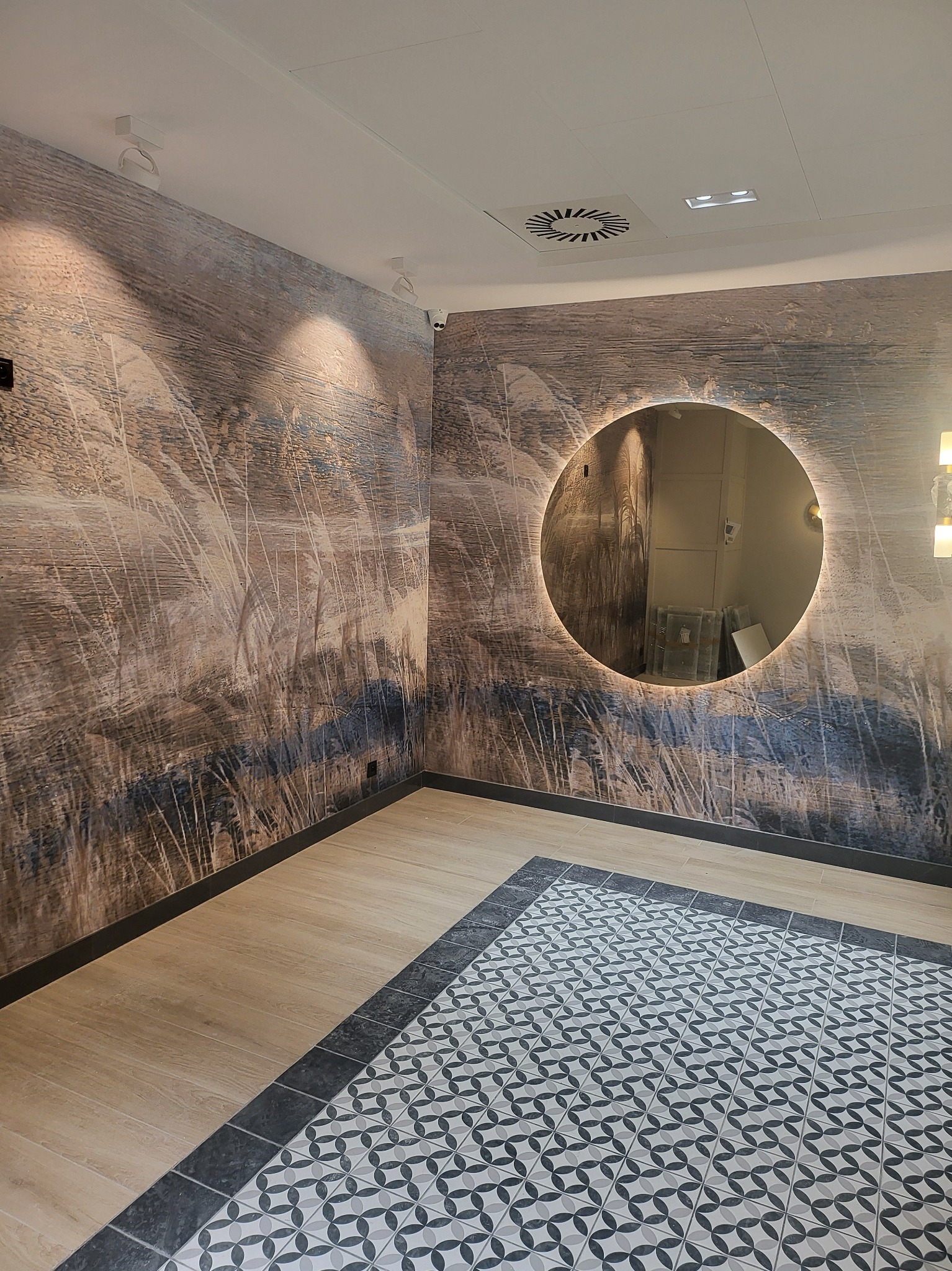Wnętrze z dekoracyjną tapetą z motywem trawy, okrągłym lustrem z podświetleniem LED i podłogą z płytek o geometrycznym wzorze.