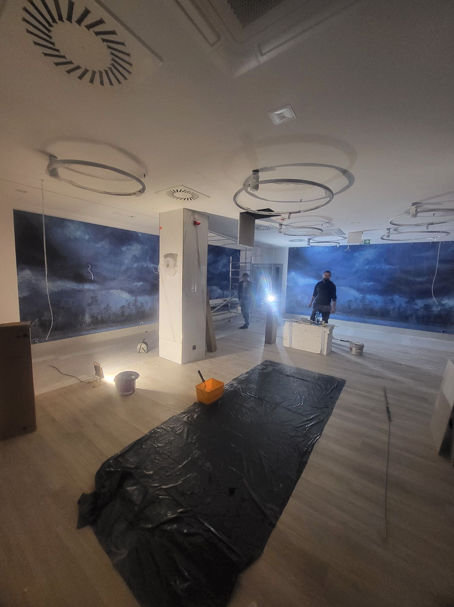 Przygotowanie do prac malarskich w pomieszczeniu z dekoracyjną tapetą: rozłożona folia ochronna na podłodze, widoczne narzędzia, pracownik w tle, oświetlenie budowlane.