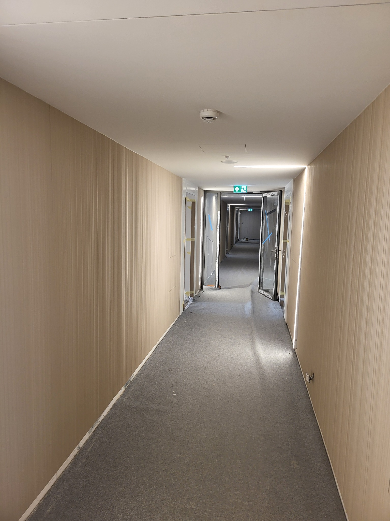 Długi korytarz z tapetowanymi ścianami w odcieniach beżu, szara wykładzina, białe listwy przypodłogowe i sufit, widoczne drzwi na końcu korytarza i oświetlenie LED wzdłuż ścian.