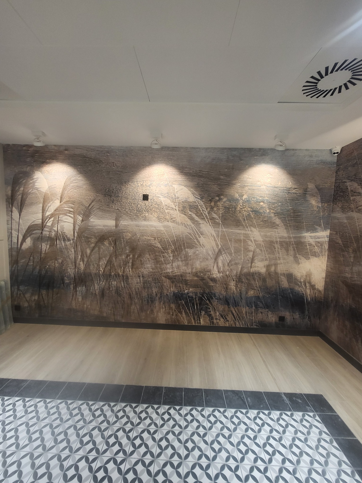 Pokój po tapetowaniu z motywem trawy, oświetlony trzema punktowymi lampami, z fragmentem podłogi wyłożonej wzorzystymi płytkami.