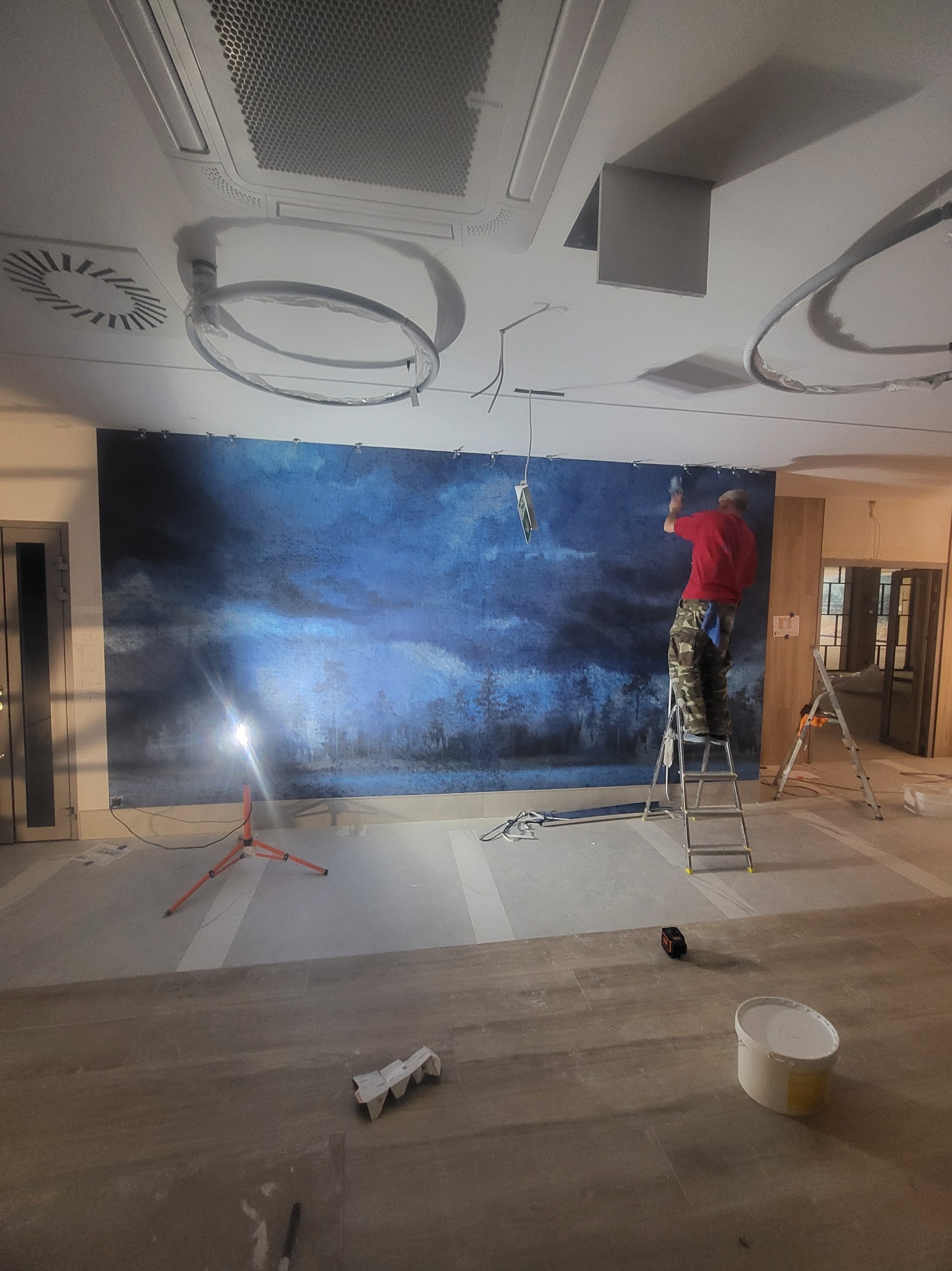 Mężczyzna na drabinie mocuje duży, dekoracyjny panel ścienny z motywem nocnego krajobrazu, oświetlony lampą budowlaną; widoczne elementy instalacji sufitowej.