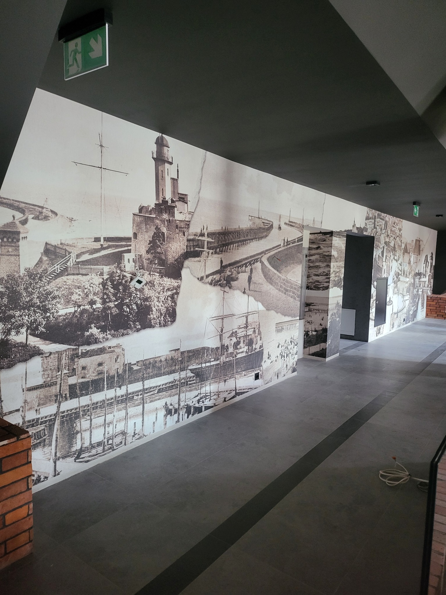 Długa ściana w korytarzu pokryta fototapetą w stylu retro z motywami portowymi i miejskimi, z widoczną fakturą i delikatnymi uszkodzeniami imitującymi starą fotografię.