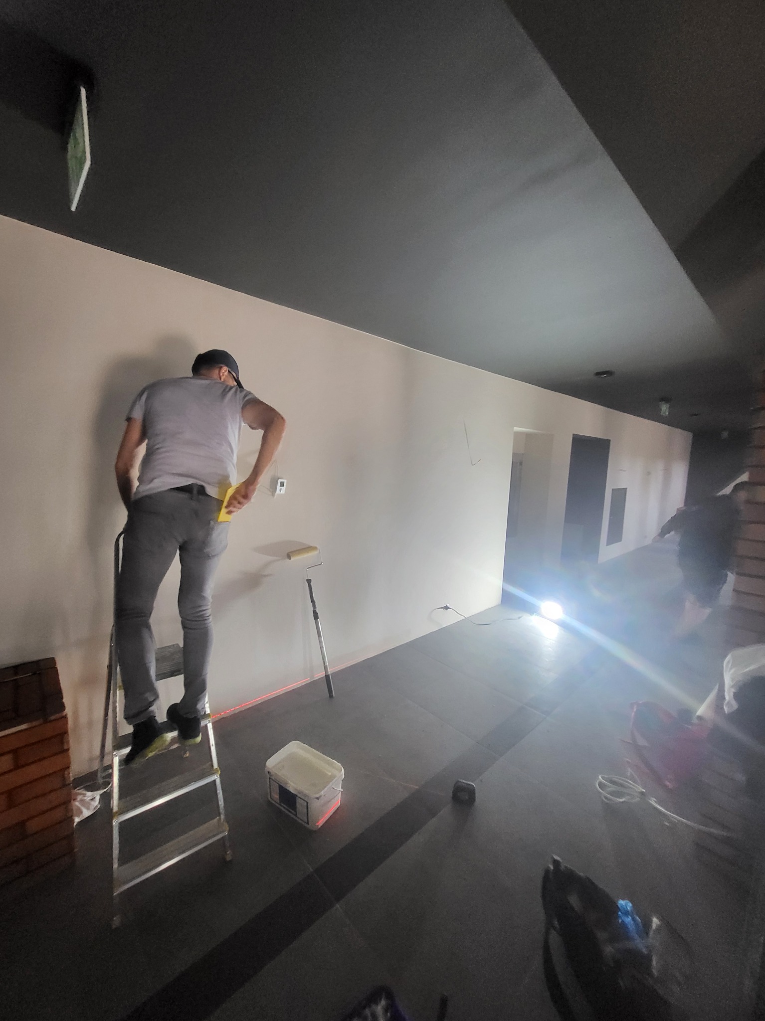 Mężczyzna w szarych spodniach i koszulce, stojący na aluminiowej drabinie, wygładza ścianę żółtą szpachelką; w tle widoczny wałek malarski, wiaderko z masą i laserowy poziom na ścianie.