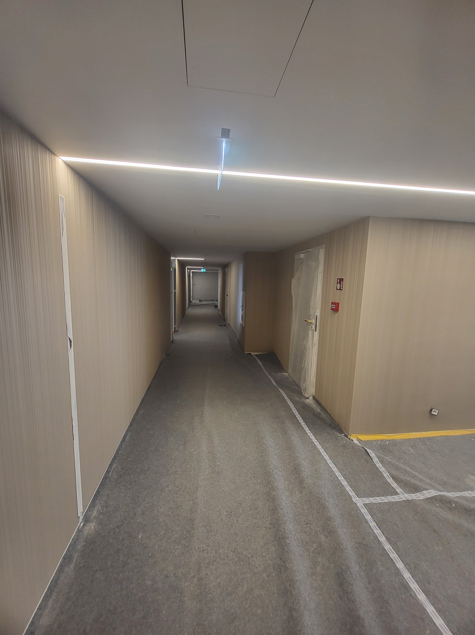 Korytarz w trakcie prac wykończeniowych; ściany pokryte tapetą o delikatnej strukturze, podłoga zabezpieczona folią ochronną, widoczne taśmy malarskie przy listwach przypodłogowych.