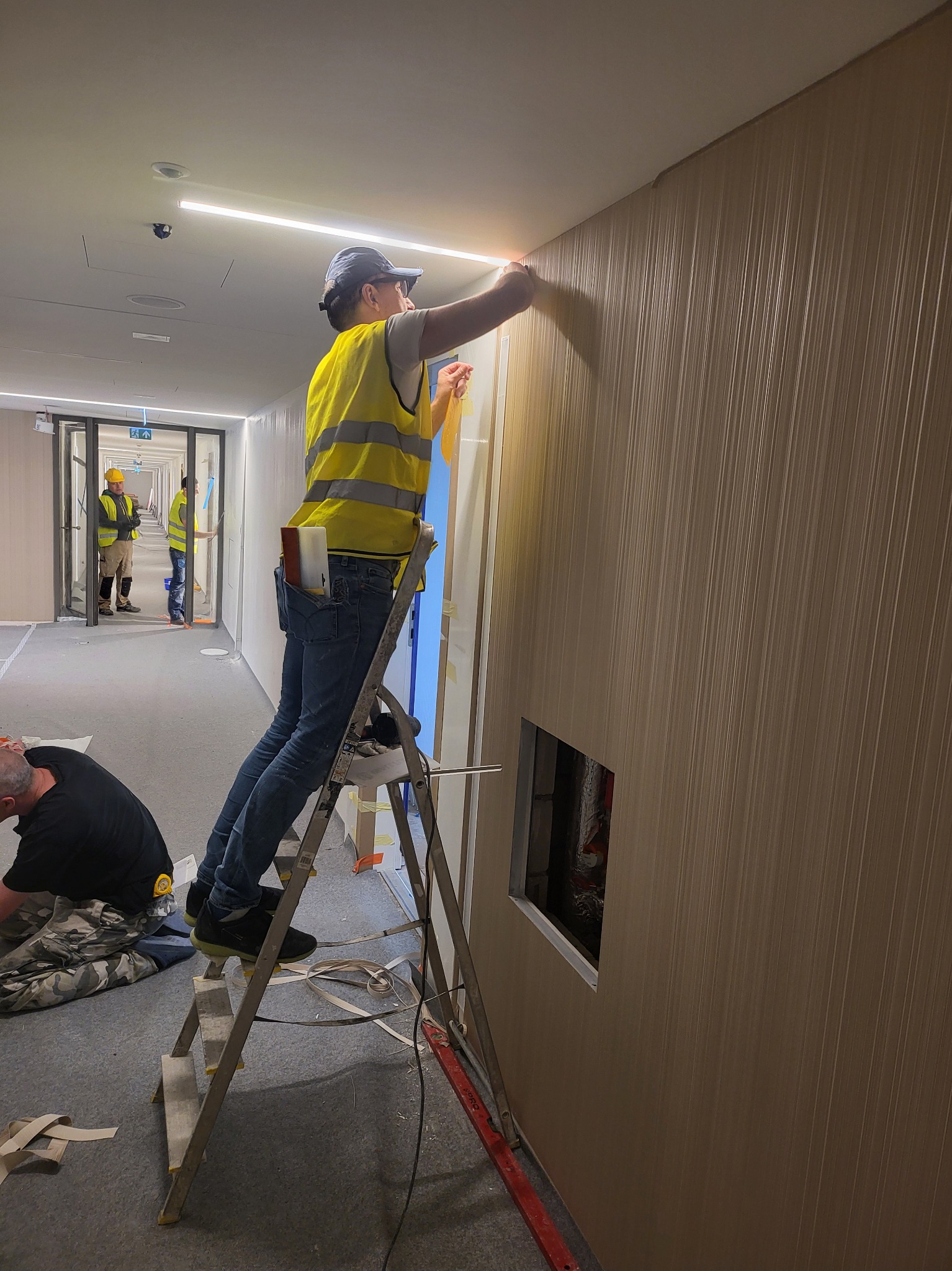Pracownik w kamizelce odblaskowej na drabinie, przykłada tapetę z pionowymi paskami do ściany w korytarzu biurowym, widoczny fragment otworu w ścianie i współpracownicy w oddali.